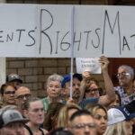A crowd sits at a public meeting. One person holds a large sign reading PARENTS RIGHTS MATTER in bold letters above their head. Other attendees look on attentively.