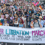 Transgender activists at a march. Pictured: a diverse crowd marches, holding a banner reading Queer Liberation March: Breaking the Chains of War and Oppression for Trans and Immigrant Rights amid colorful signs and pride flags.