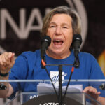 AFT's Randi Weingarten is influencing AI through corporate partnerships. Pictured: a person wearing a blue shirt speaks passionately at a podium with two microphones, gesturing with one hand. A blurred logo and flames are visible in the background.
