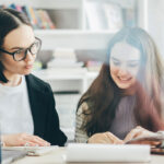 The American School Counselor Association has admitted that neurodiversity is a term for critical race theory and queer theory. Pictured: Two women sit at a table with books and a laptop, smiling as they look at papers together in a bright, cozy room with shelves of books in the background.