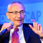 The Center for American Progress helped launch Faith in Public Life, which was meant to be an "organizational anchor for the Religious Left." Pictured: John Podesta, a man in a suit and red tie speaks into a microphone, gesturing with his hand. A blue background displays blurred text and numbers.
