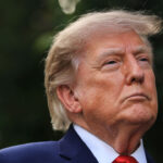 President Donald Trump, a man with light skin and blond hair wearing a navy suit, white shirt, and red tie looks upwards with a serious expression. The background is blurred greenery.