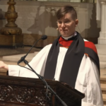 Rev. Flouish Klink claims Jesus is trans. Pictured: a person in religious robes stands at a carved wooden pulpit, speaking into a microphone, with one hand gesturing outward inside a large, ornate church.