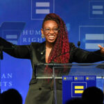 Human Rights Campaign President Kelley Robinson, a person with long red braids and glasses smiles and gestures with open arms while standing at a podium, against a blue Human Rights Campaign backdrop.