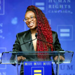 Human Rights Campaign President Kelley Robinson compared ICE to lynch mobs. Pictured: a person with long red braids, glasses, and a dark blazer speaks at a podium with a Human Rights Campaign logo against a blue background.