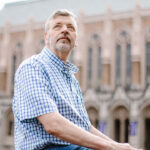 University of Washington professor Stuart Reges, who got disciplined over a land acknowledgment. Pictured: Reges, an older man in a blue plaid shirt sits outdoors, looking upwards thoughtfully. A large, historic building with arched windows and detailed stonework is in the blurred background.