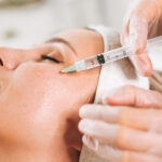 A person receiving a cosmetic facial injection near the cheekbone, with a gloved professional administering the treatment. The person has their eyes closed and is lying back with a towel around their head.