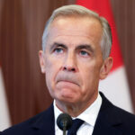 Mark Carney in a suit stands in front of two red and white flags, looking serious, with a microphone visible in front of him.