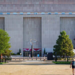 A person with a backpack walks toward the Smithsonian's Museum of American History, a large, modern building with tall columns. Trees line the path, and a metallic sculpture stands near the entrance.
