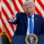 Donald Trump in a suit speaks at a podium with the U.S. presidential seal, pointing forward as he addresses matters related to Iran. American flags hang in the background.