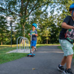 Two boys wearing helmets ride scooters on a paved path in a park surrounded by trees and grass on a sunny day.