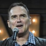 A man wearing a black jacket and checkered shirt speaks into a microphone, standing in front of a background with large, illuminated letters.