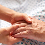 Feminine hands hold the hand of an elderly patient in white hospital gown with blue dots.