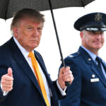 Donald Trump in a suit holds an umbrella and gives a thumbs up, standing next to a uniformed officer outdoors.
