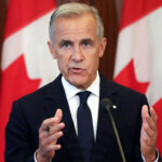 Mark Carney in a dark suit and tie speaks in front of several large Canadian flags, gesturing with his hands during a press conference or public address about tariffs.
