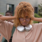 A person from the younger generation with curly hair, glasses, and headphones around their neck covers their ears and looks upset. Two people stand in the background outdoors.