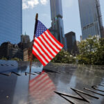 An American flag stands on the engraved names at the 9/11 Memorial in New York City, with tall buildings and trees in the background on a sunny day.