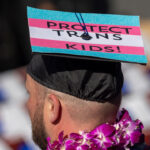 A person wearing a graduation cap decorated with the trans pride colors and the words Protect Trans Kids! stands among a crowd, with a purple flower lei around their neck. Teachers are being taught to indoctrinate according to a new study.