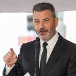 A man in a black suit and tie speaks into a microphone, gesturing with his right hand. Reminiscent of Kimmel’s charismatic stage presence, he has short, dark hair and a trimmed beard. The background is blurred, suggesting an outdoor event.