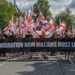 A group of people march holding flags and a banner that reads, REMIGRATION NOW MILLIONS MUST LEAVE, highlighting their stance on immigration, with a police van beside them and trees in the background.