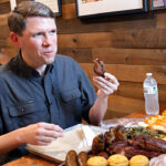 James Talarico sits at a wooden table, eating barbecue with sides like cornbread, greens, and macaroni. He holds a rib. A water bottle, napkins, and sauces are also on the table. Another person sits in the background.