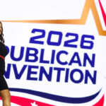 Monica De La Cruz in a black dress waves while standing in front of a large sign that says 2026 Republican Convention with a star graphic.