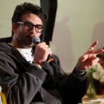 A piker wearing glasses and a dark jacket speaks into a microphone, gesturing with two fingers. There are drinks and flowers on the table in the background, and another person is faintly visible.