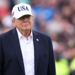 Donald Trump wearing a white USA cap, dark jacket, and white shirt stands outdoors with a blurred crowd in the background.