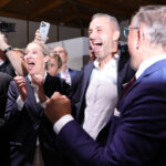 A group of people from the AfD in business attire cheer and celebrate together indoors. Some are smiling and shouting, while others take photos. The atmosphere is lively and joyful.