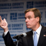 Mark Warner in a suit gestures with his right hand while speaking at a podium. The background displays the Fairfax County Chamber of Commerce logo and text repeatedly.