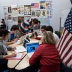 Students sit at tables in a classroom, using laptops for schoolwork. Educational posters and maps—including references to Western civilization—decorate the walls. An American flag is in the foreground. Small groups work together, engaging with their computers and each other.
