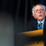 An older man with glasses and gray hair speaks passionately at a podium, wearing a suit and tie, with dark curtains in the background. The podium has the partially visible word ASSEMBLY..