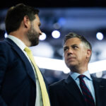 JD Vance and Jim Banks in suits stand close together, facing each other and talking, with a blurred crowd and bright lights in the background.