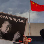 A person holds a #FreeJimmyLai sign with a black-and-white photo of a man. In the background, a Chinese flag flies above a building under a cloudy sky.