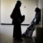 Two people in shadowed clothing stand and sit facing each other in a dimly lit, mostly empty hallway with light streaming through a nearby window.