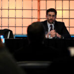 Hasan Piker in a suit speaks into a microphone at a table on stage, facing an audience. The background is illuminated with orange grid-like lights, creating a dynamic atmosphere.