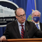 Jeff Clark in a suit and glasses speaks at a podium with the Department of Justice seal behind him. Another man in a suit and face mask stands in the background near U.S. flags.