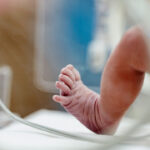 Close-up of a newborn baby’s foot inside an incubator, with medical tubes visible in the blurred foreground. The baby’s toes are curled, highlighting the fragility and care required in a hospital or neonatal unit—a poignant reminder of the complex decisions surrounding birth and abortion.