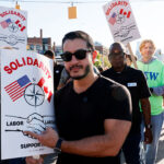 A group of people march on a street holding Solidarity signs with US and Canadian flags, a compass, and shaking hands, supporting labor unions and advocating for fair election practices. Abdul El-Sayed in sunglasses holds a sign in the front.