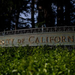 A metal sign reads University of California in gold letters, partially obscured by green foliage, with trees and a dark background behind it.