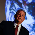 Al Gore in a suit speaks while holding a remote, with a large image of Earth projected behind him.