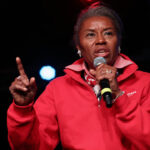Winsome Earle-Sears with graying hair, wearing a red quarter-zip sweatshirt, speaks into a microphone on stage. She is pointing her index finger up, and colorful stage lights glow in the dark background, capturing a commanding presence reminiscent of Earle-Sears.