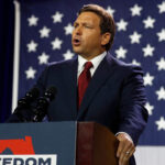 Ron DeSantis in a suit speaks at a podium labeled FREEDOM with a Florida outline, in front of a large American flag, emphasizing the importance of election integrity.