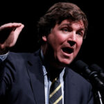 Tucker Carlson in a suit and striped tie speaks passionately into multiple microphones against a dark background, gesturing with his right hand raised, reminiscent of the commanding presence of Tucker Carlson during a televised broadcast.