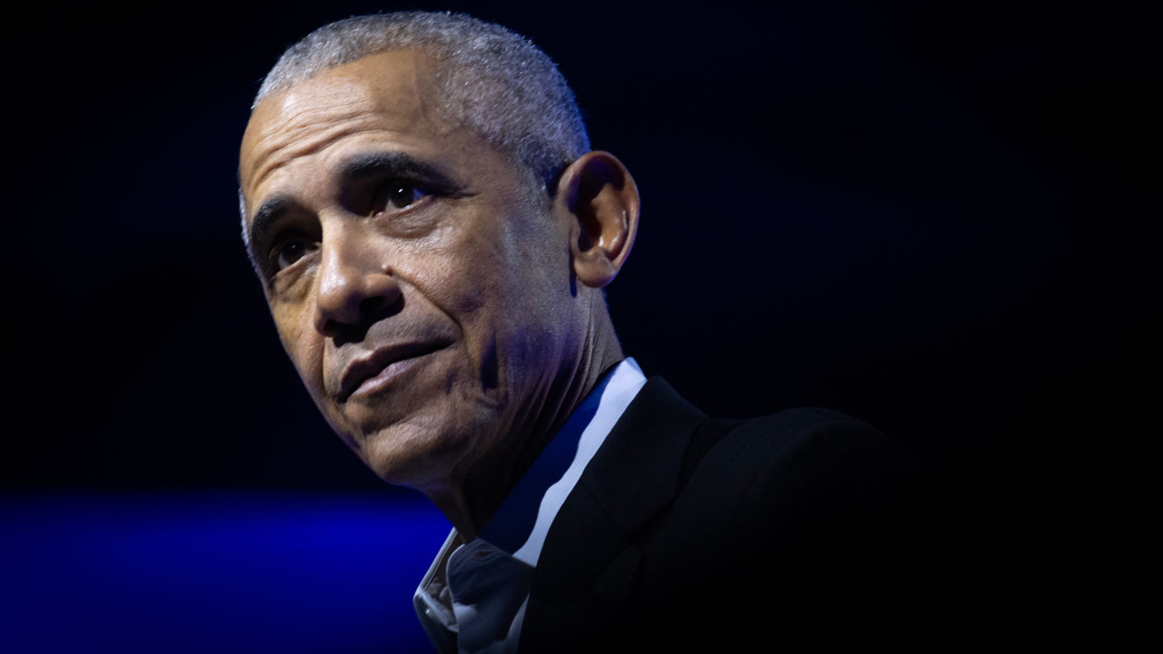 Home 3 Barack Obama with short gray hair wearing a dark suit and white shirt looks thoughtful against a dark blue background with dramatic lighting.