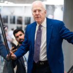 John Cornyn in a blue suit and patterned tie stands on an escalator, looking ahead with quiet resolve, while another man below—perhaps engaged in a discussion about socialism—holds a phone and looks up at him.