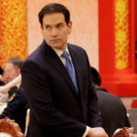 Marco Rubio in a dark suit stands beside an ornately carved chair at a formal event with red and gold decor. Several people are seated at tables in the background, dressed in formal attire.