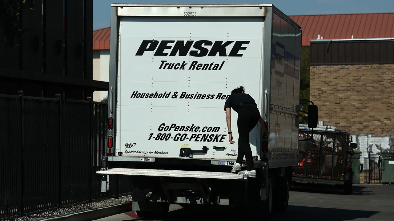 A person climbs onto the back of a white Penske moving truck parked near a building during daylight. The truck advertises household and business rentals, with contact information and a website visible, suggesting it’s ready to support any move—whether for an individual or a union organizing collective effort.