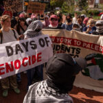 A group of protesters hold signs, including one reading 700 DAYS OF GENOCIDE in large letters, and another that says STUDENTS FOR JUSTICE IN PALESTINE. Many participants, some wearing keffiyehs stand closely together outdoors.