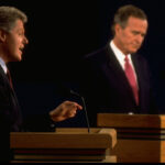 Two men in suits stand at podiums during a 1992 debate. The man on the left gestures while speaking; the man on the right looks downward, listening. Both are in front of a dark background.